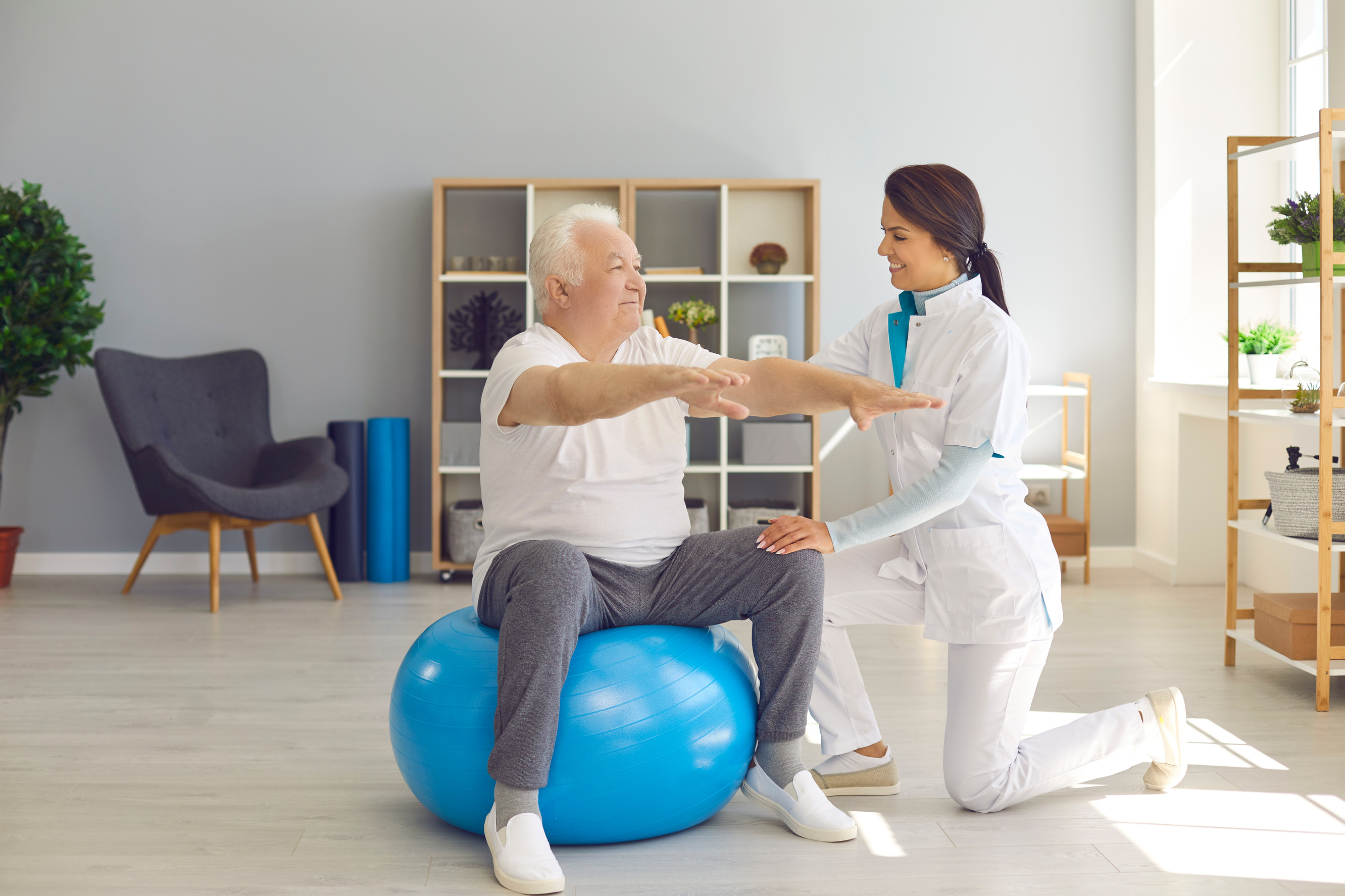 Smiling Woman Chiropractor Correcting Old Man Movements during Rehabilitation Theapy on Fitness Ball